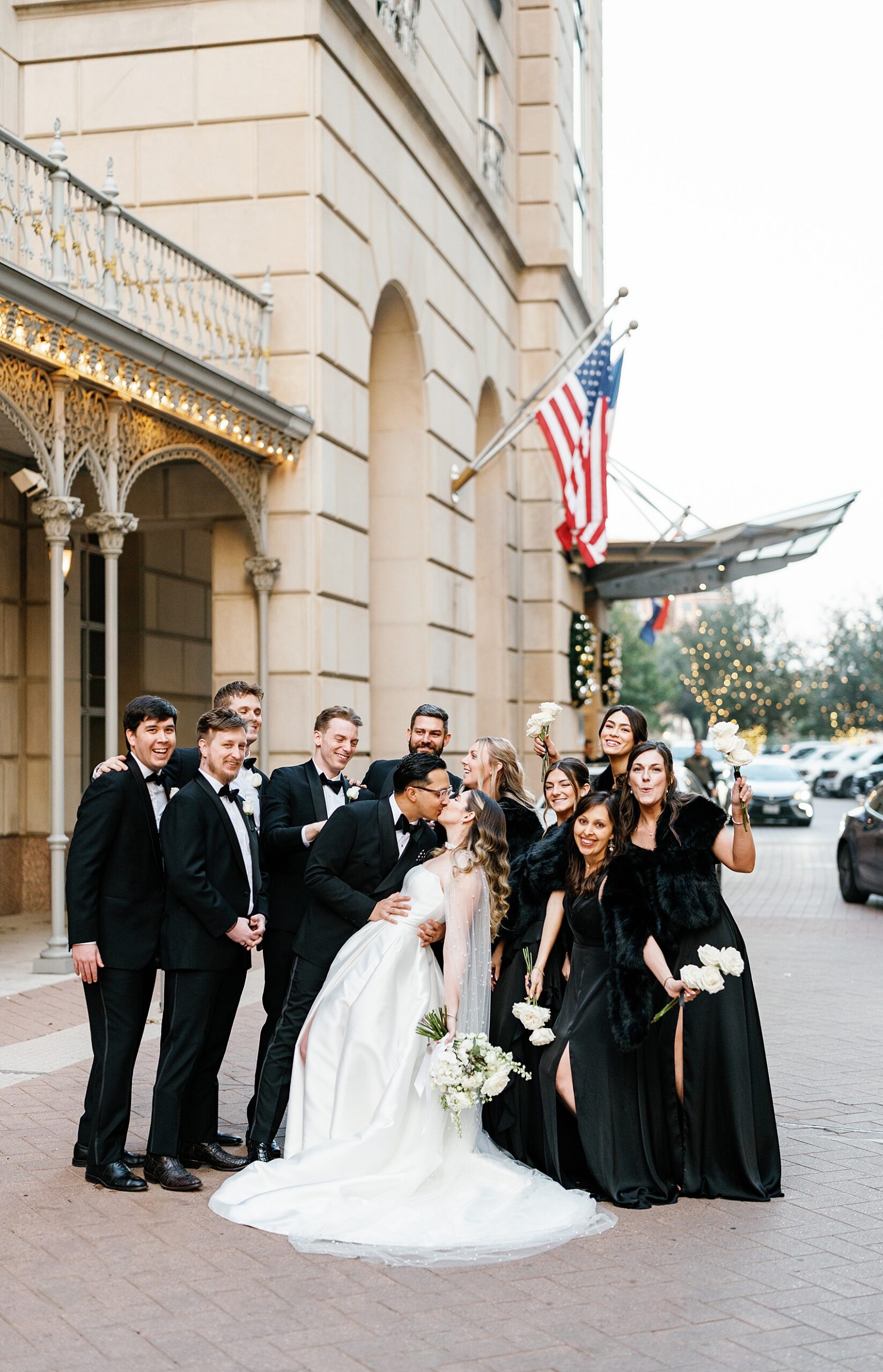 A bride and groom kiss as their wedding party in black celebrates behind them in front of the Hotel Crescent Court Wedding venue