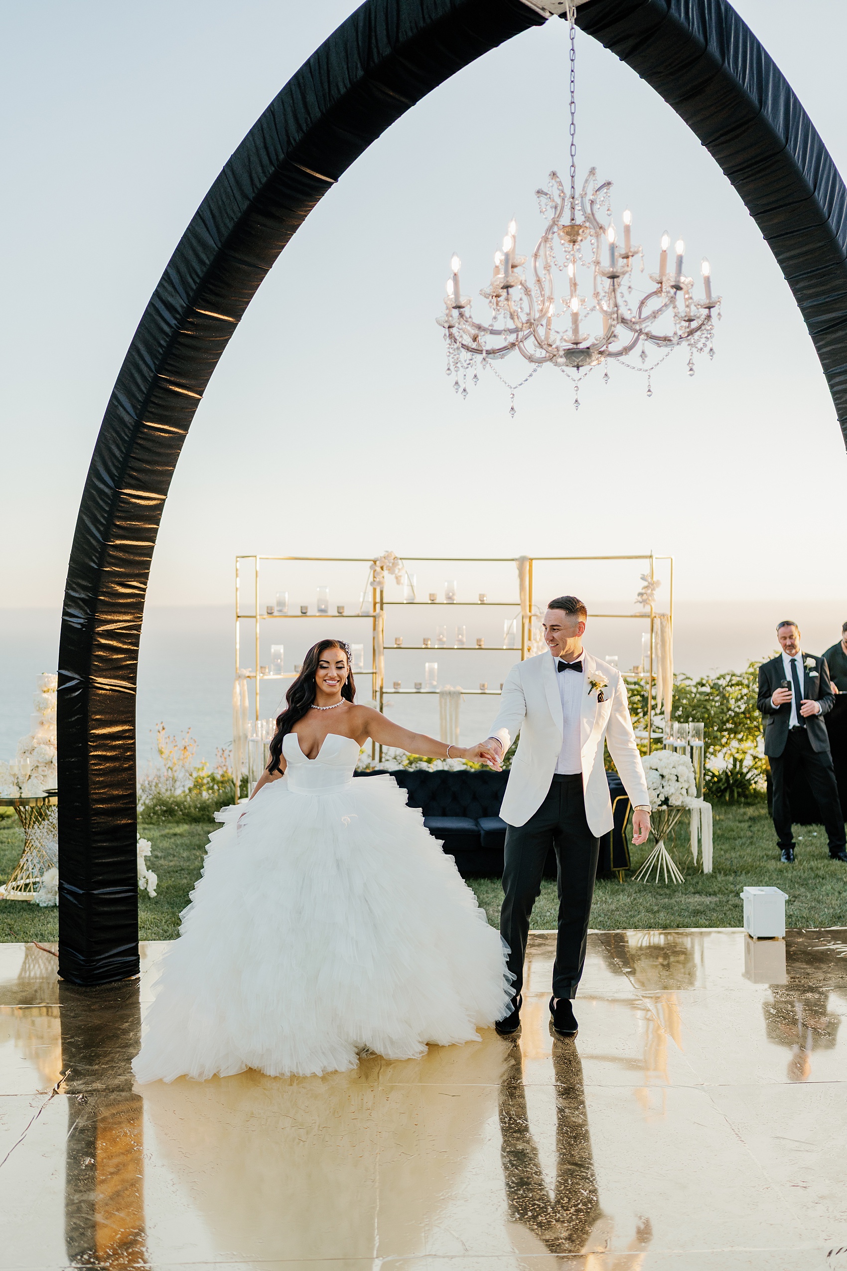 A groom leads his bride onto the outdoor dance floor for their first dance under a chandelier