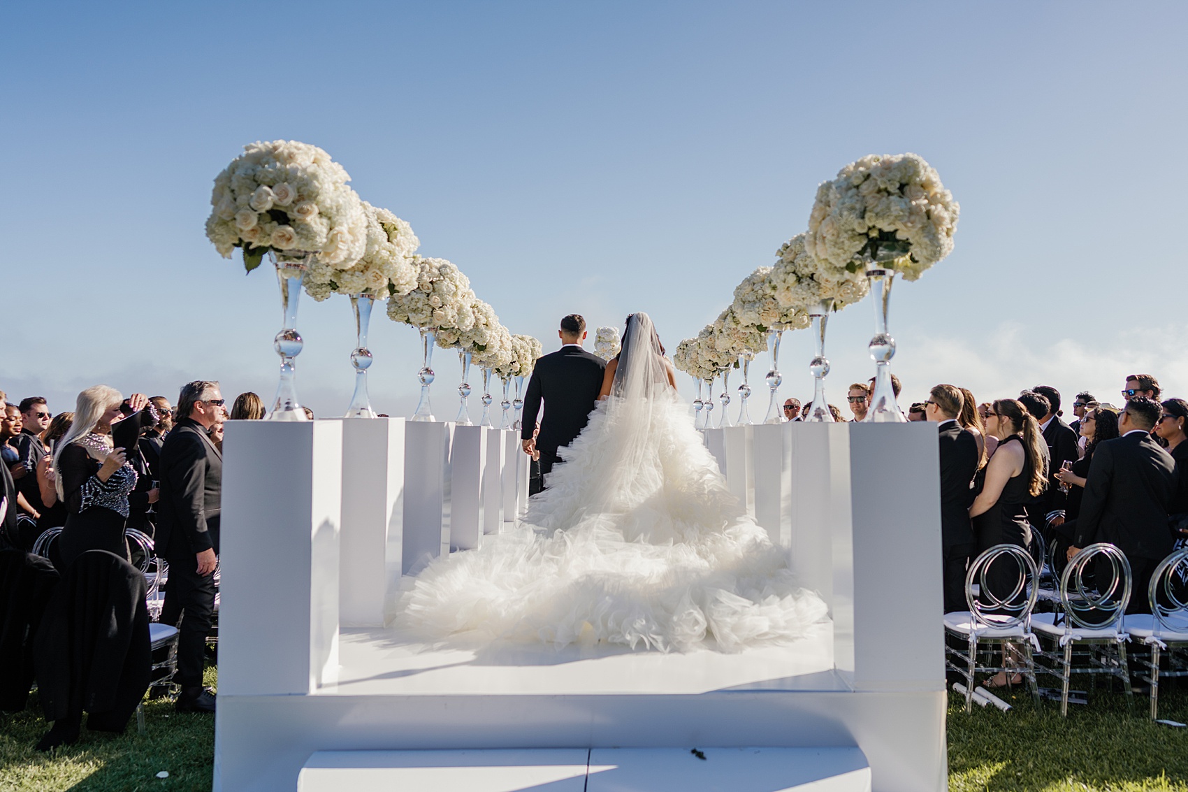A bride walks down the elevated aisle as guests look on with a long flowing tule train