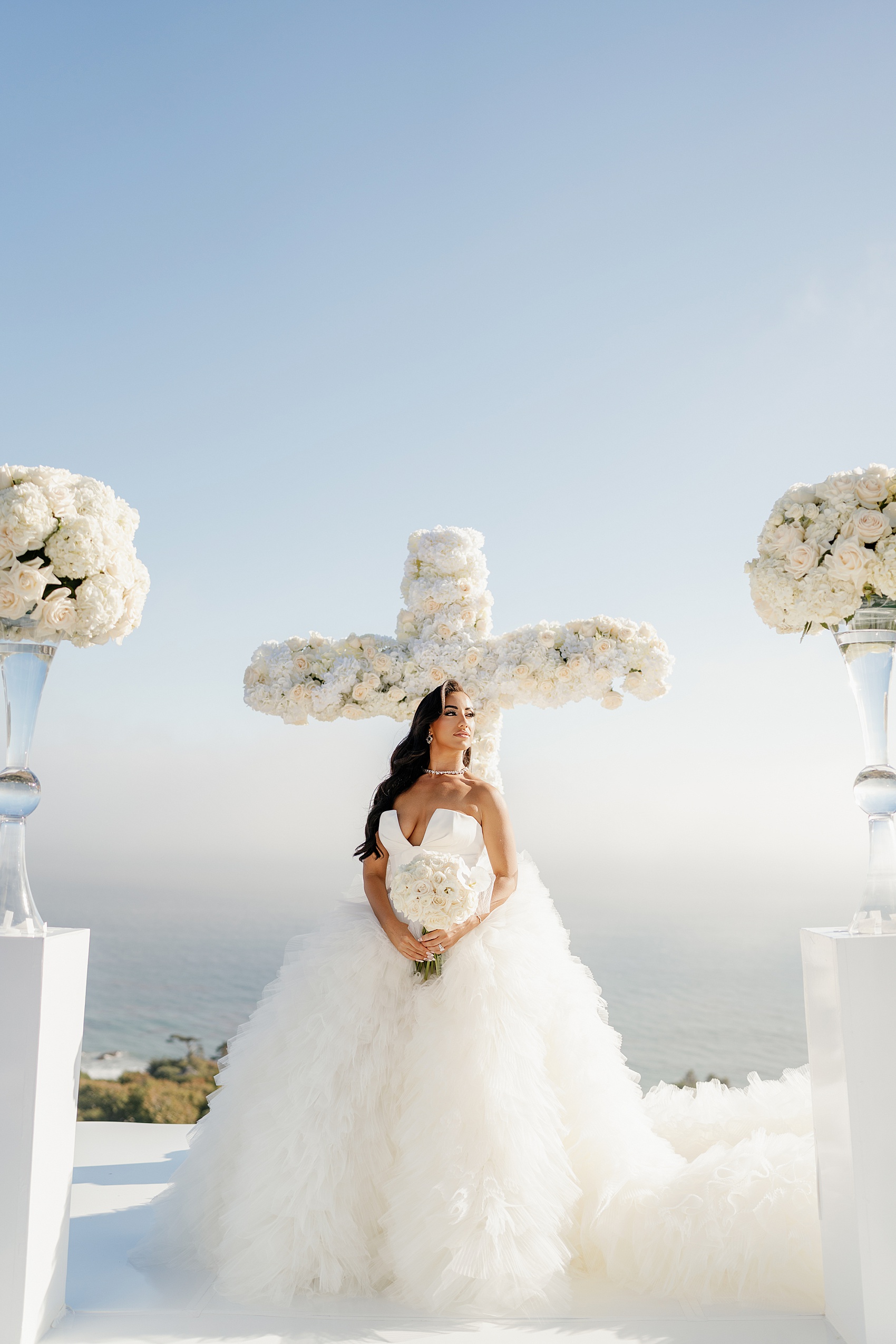 A bride stands holding her bouquet at her ceremony cross of flowers overlooking the ocean at the Malibu Sea View Estate
