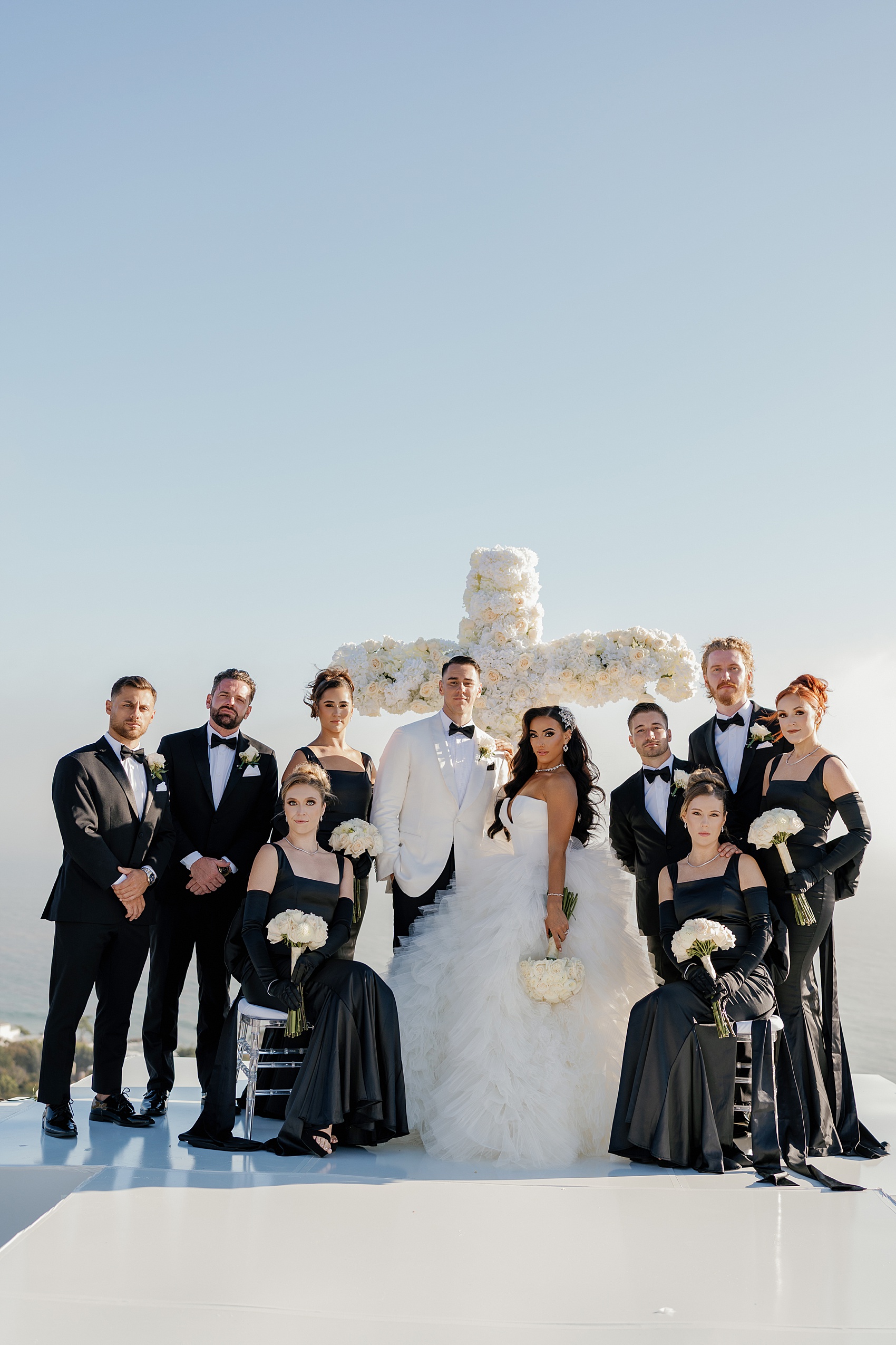 Newlyweds in white stand at their altar with their bridal party in black holding white bouquets stand stoicly