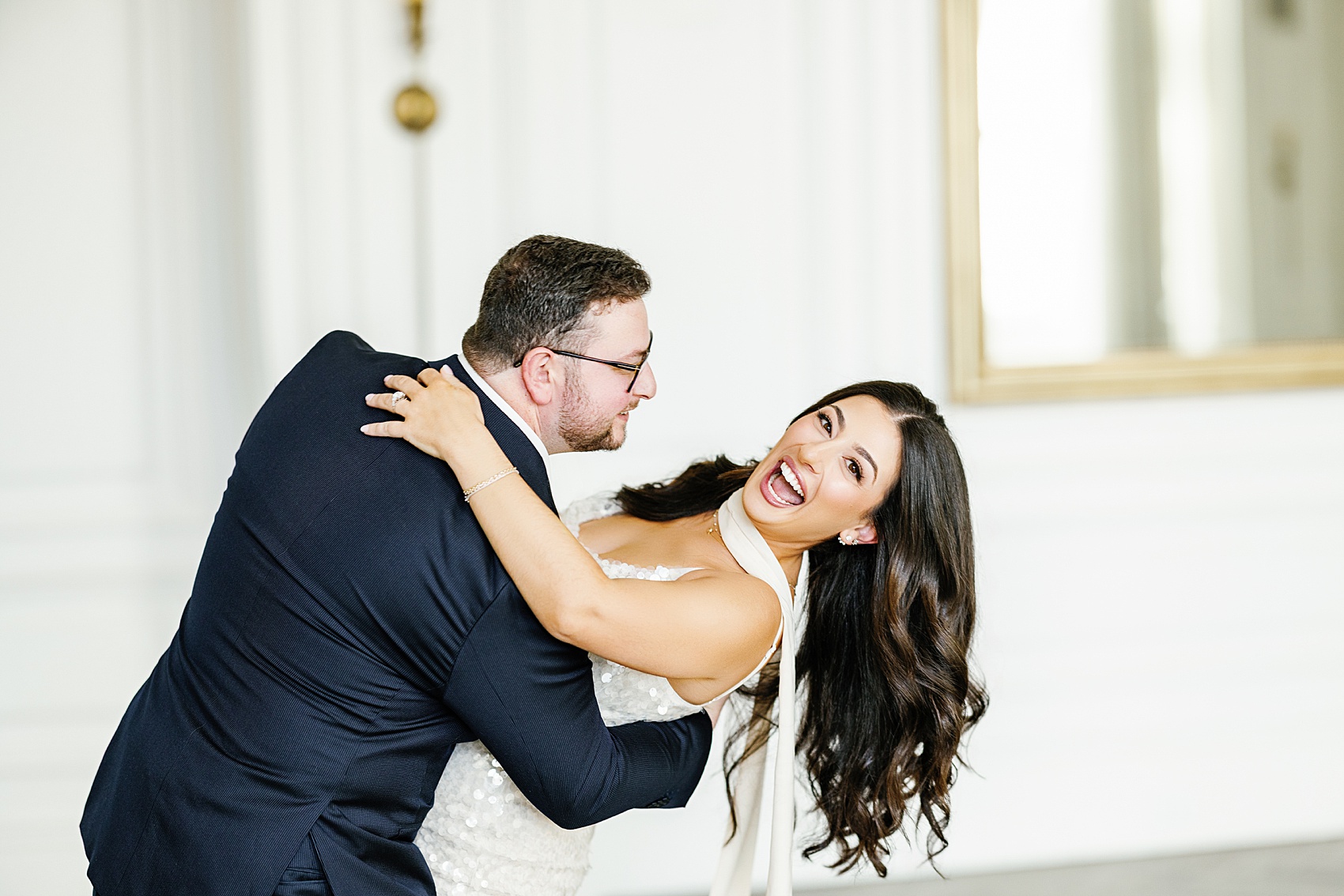 A bride laughs big while being dipped by her groom as they dance in a large white room