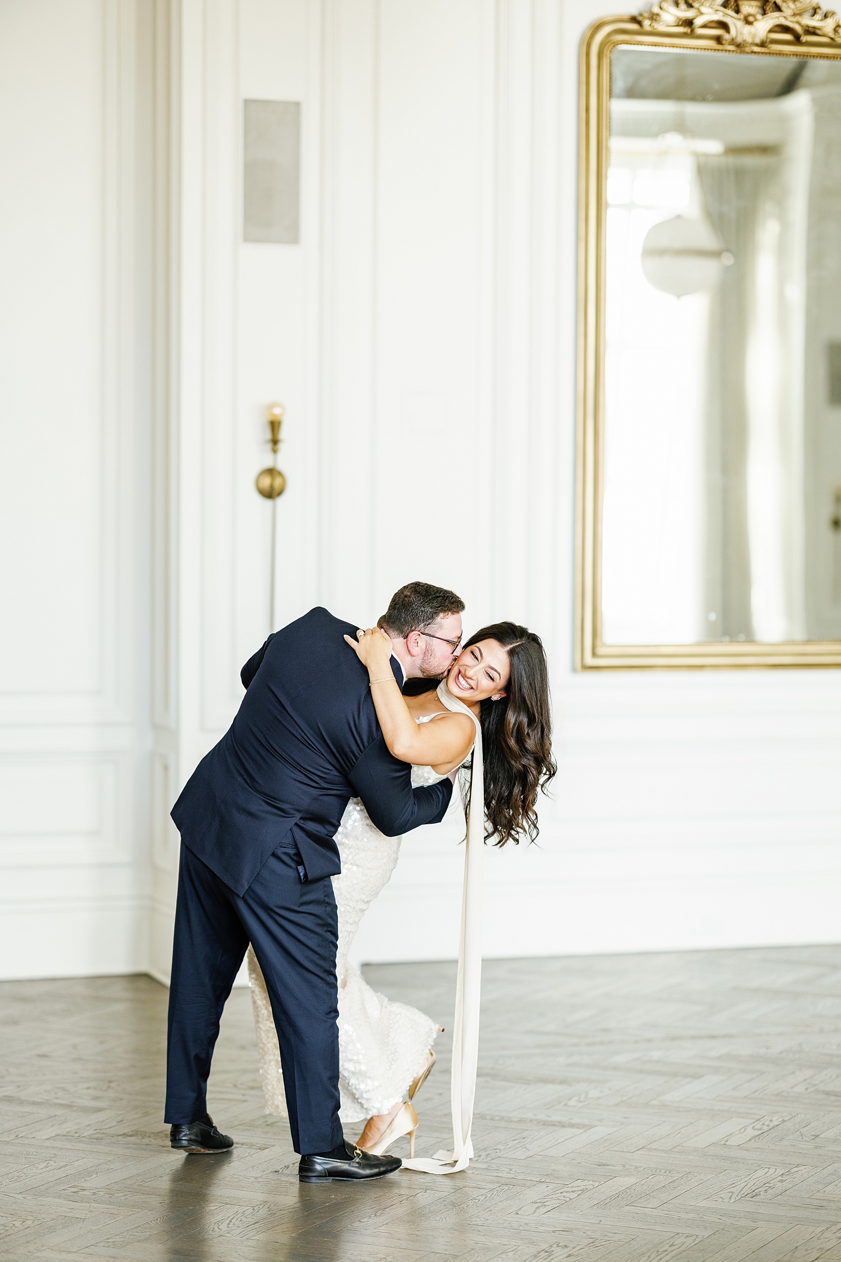 A groom dips and kisses his laughing bride in a large white room at The Adolphus Hotel wedding venue