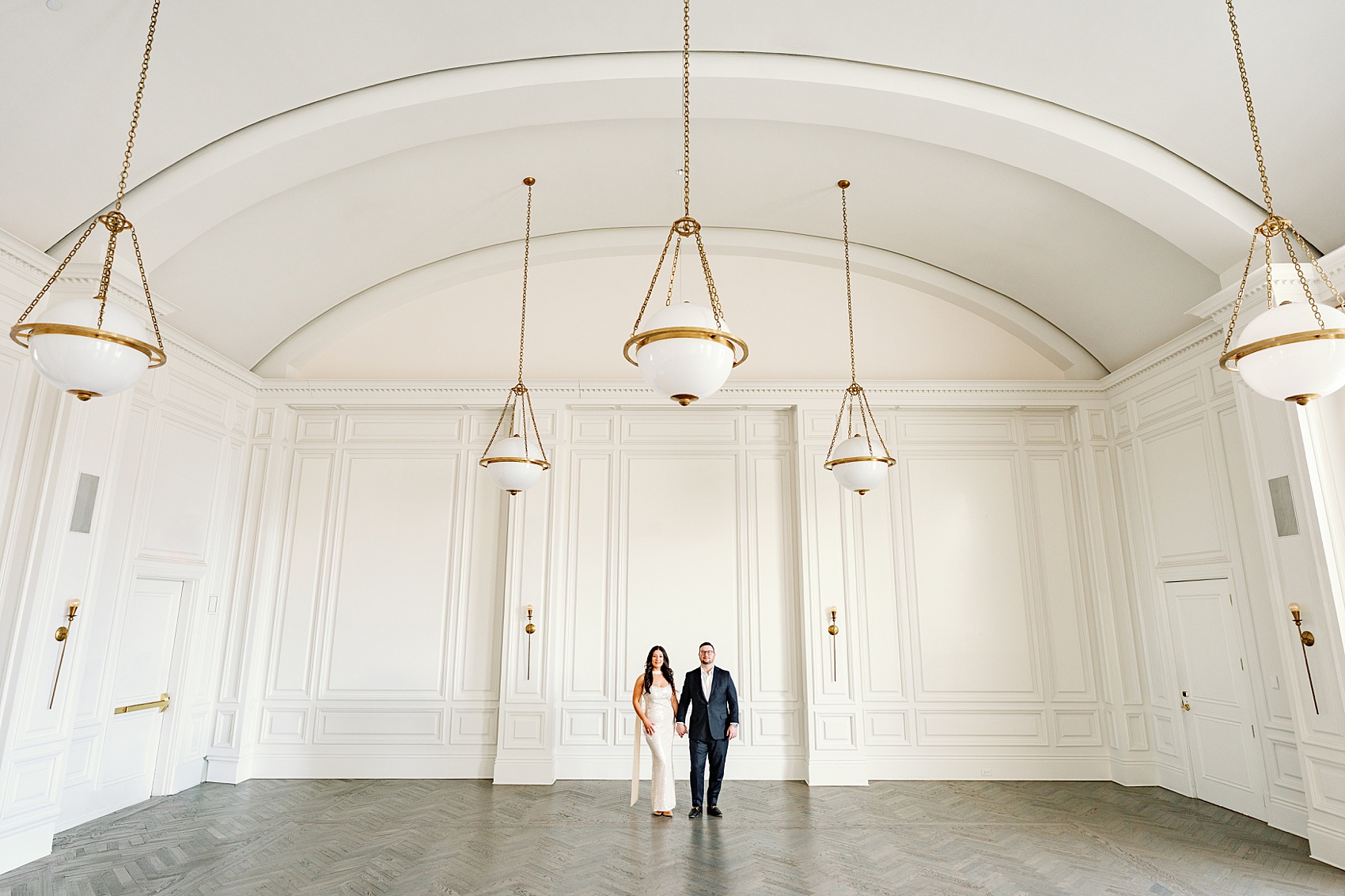 A bride walks holding hands with her groom in a black suit in a beautiful hall with chandeliers during their The Adolphus Hotel wedding