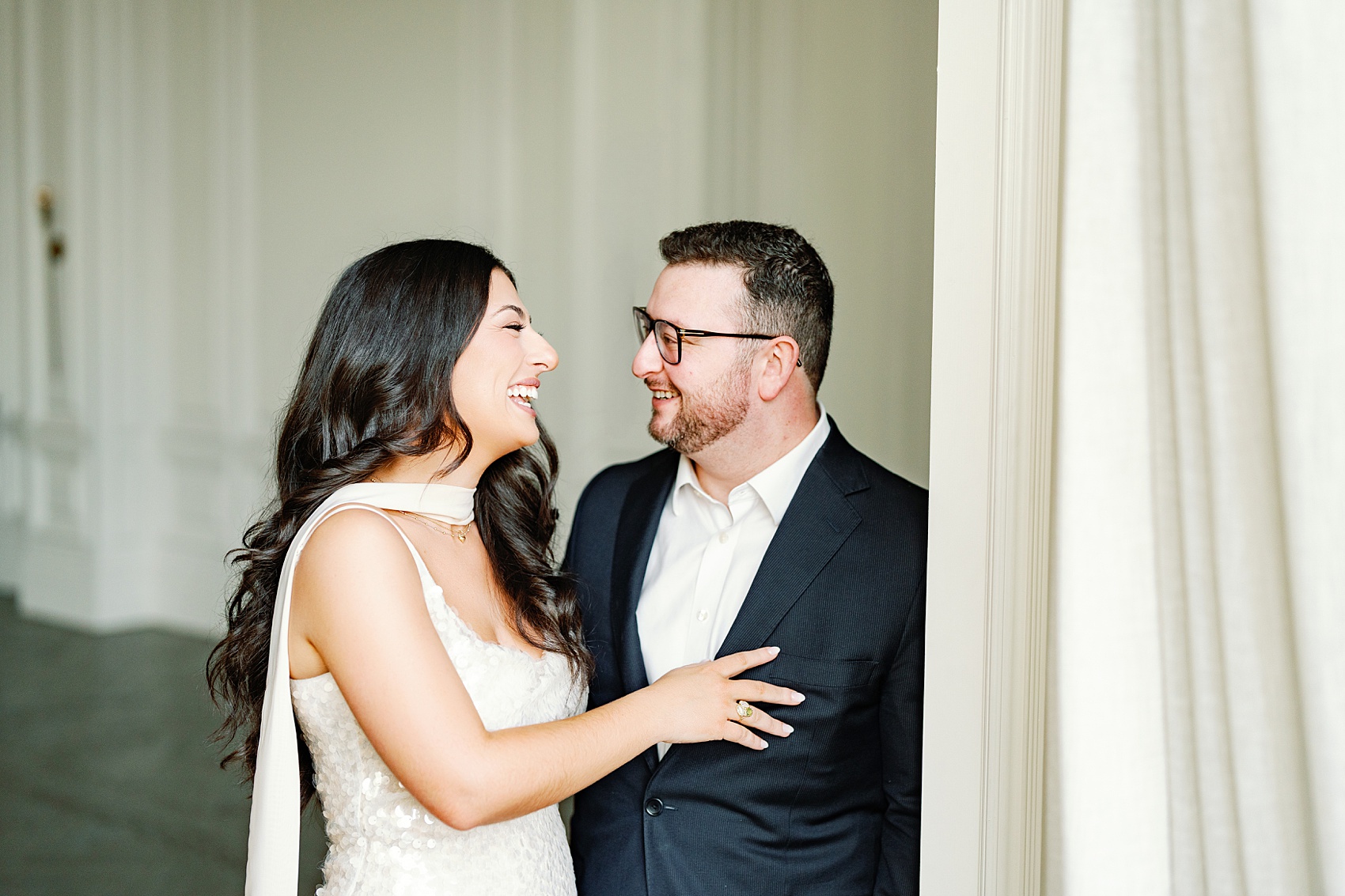 Newlyweds stand in a doorway laughing together in a black suit